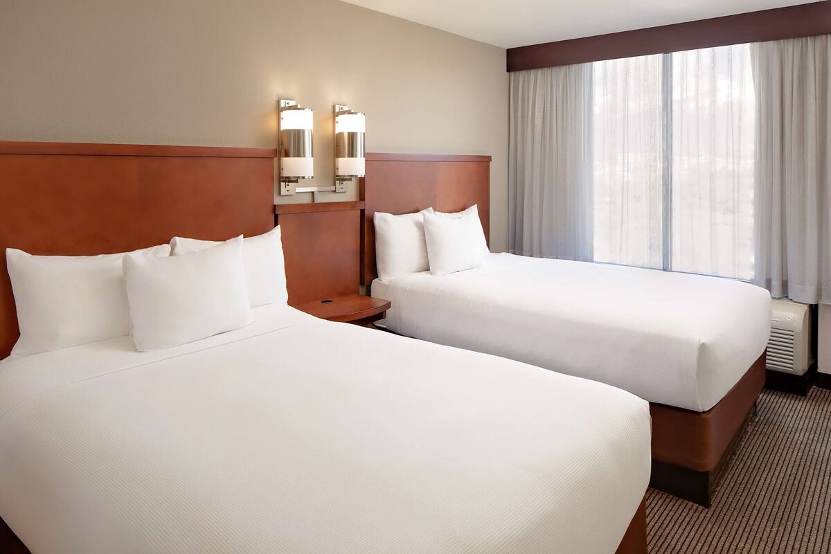 A hotel room with two beds, a wooden headboard, a nightstand, and a window with curtains.