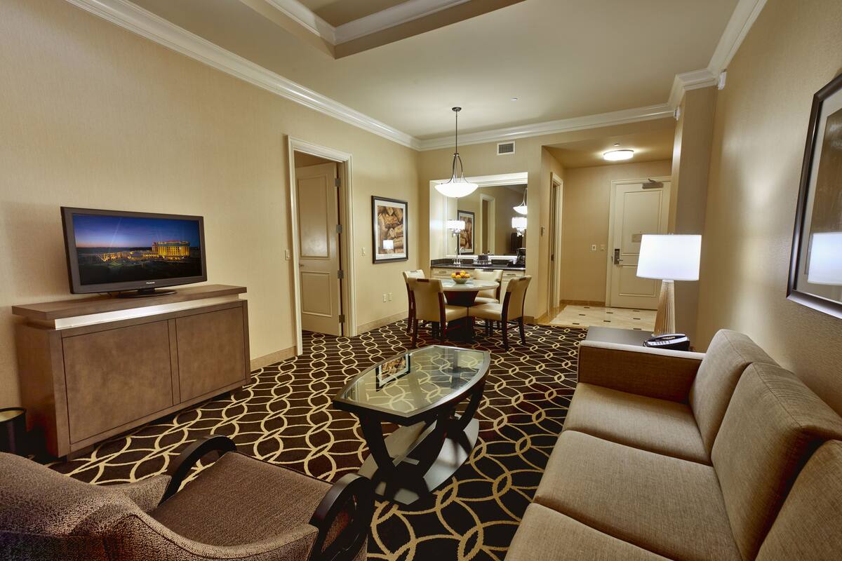 Habitación de hotel con sofá, sillón, mesa de vidrio, televisión y un área de comedor con sillas y mesa.
