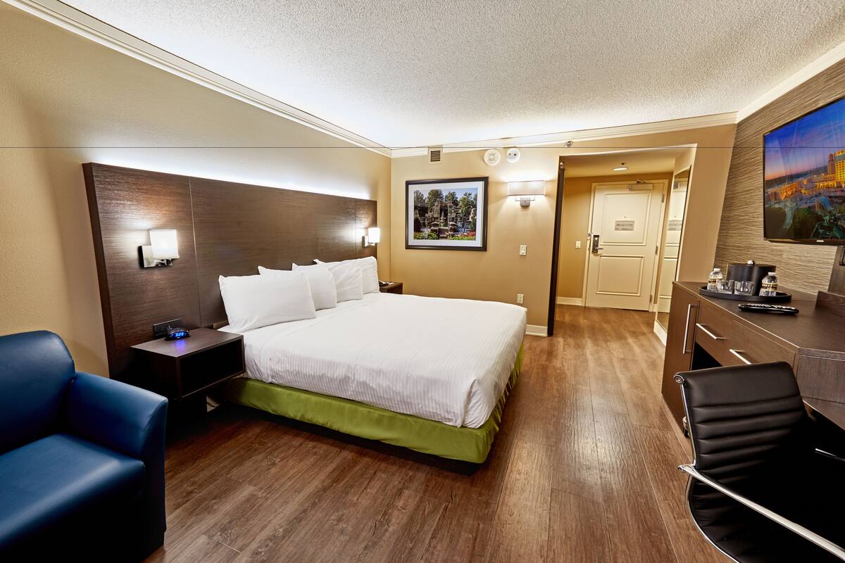 Habitación de hotel con cama grande, escritorio, silla, mesita de noche, televisor montado en la pared y puerta que da a otra habitación.