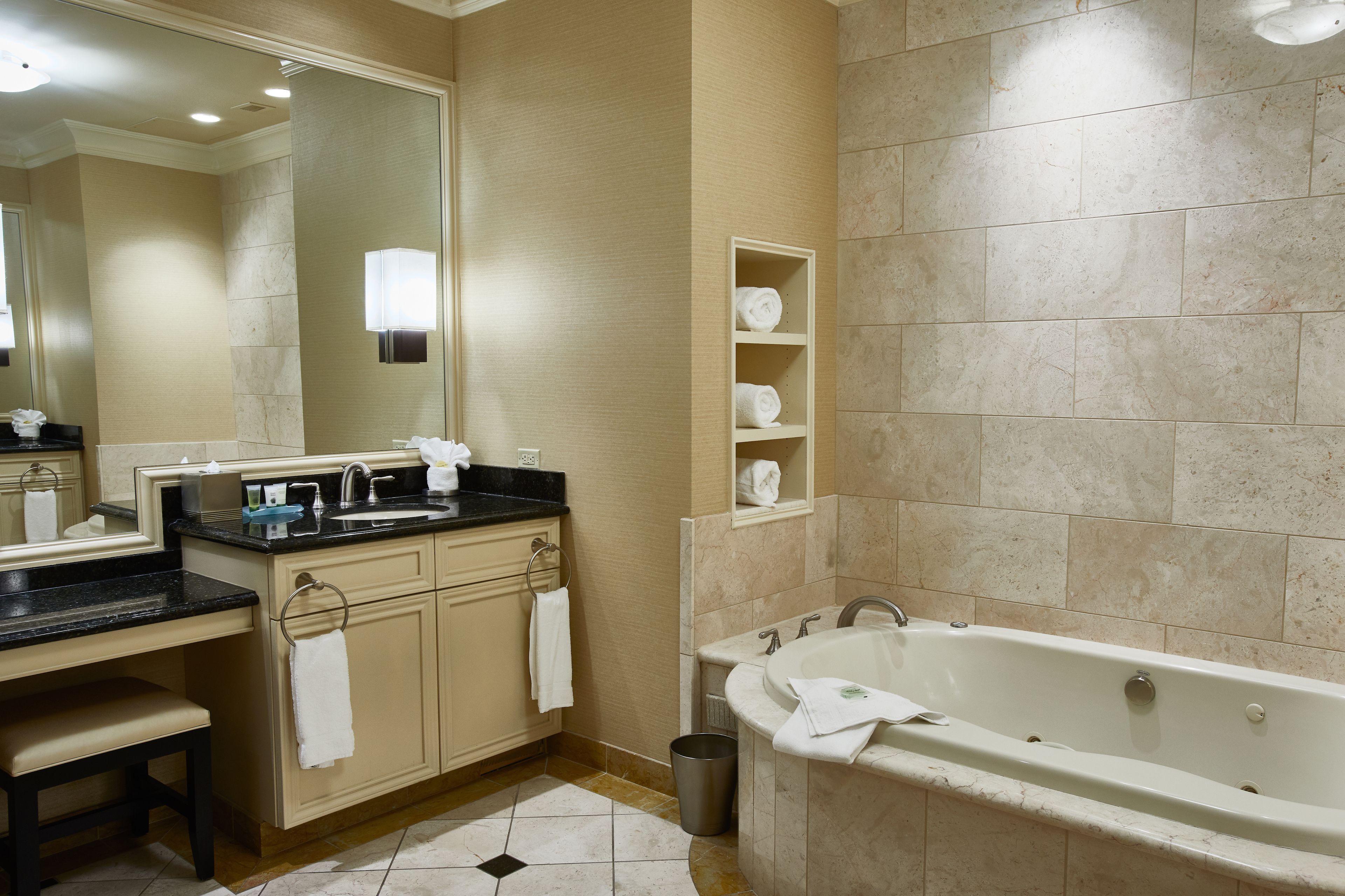 A modern bathroom with a large bathtub, a vanity with a sink, and a mirror. There are towels hanging on the wall and a trash can near the bathtub.