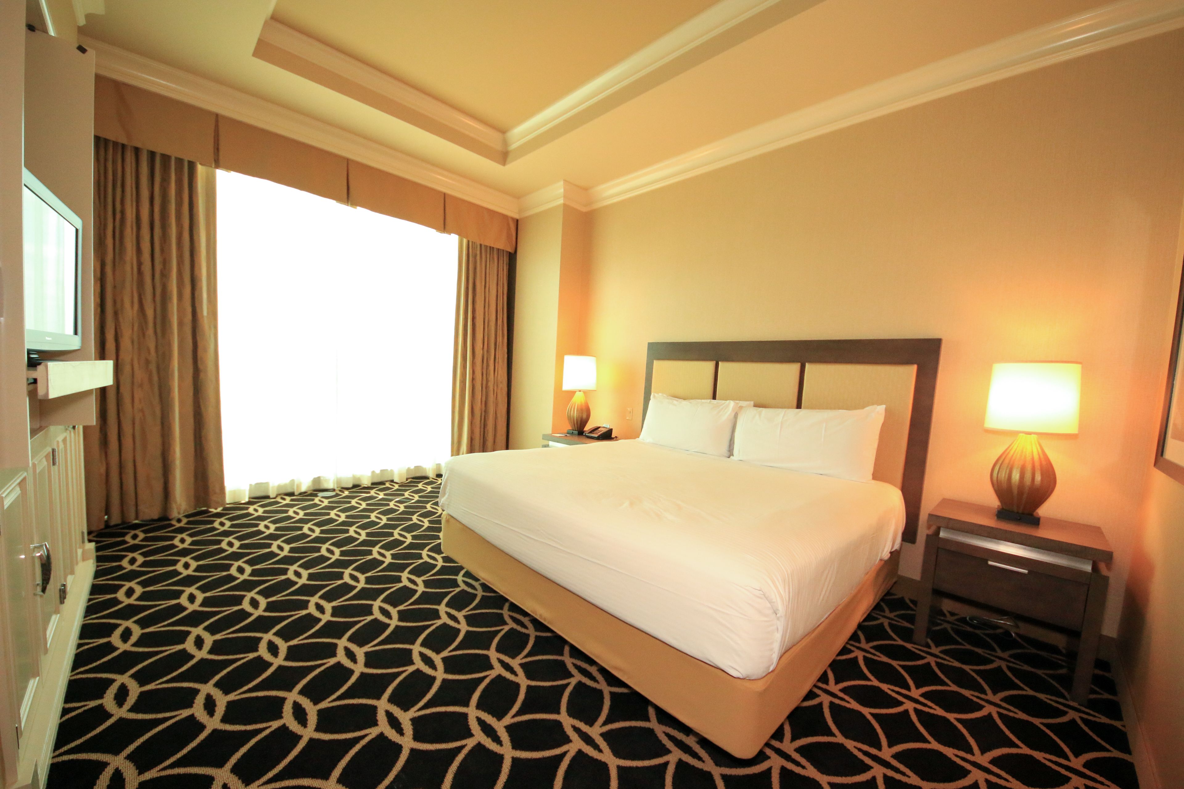 A hotel room with a large bed, two bedside tables with lamps, a television on a cabinet, and a patterned carpet.
