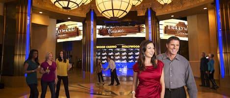 A lobby with a large screen displaying "Hollywood Casino" and "Wood Casino" signs, multiple chandeliers, and a polished floor.