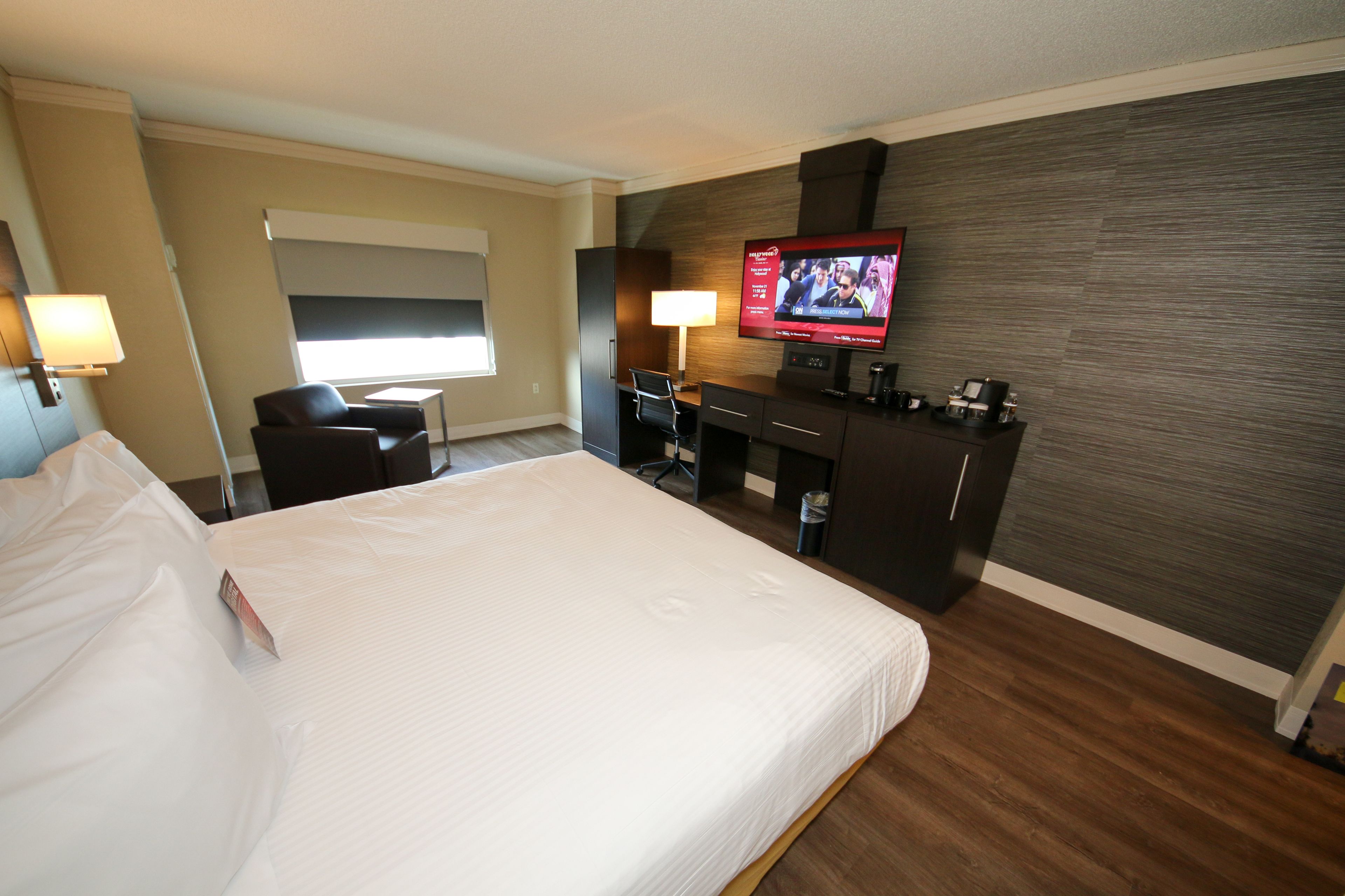 Habitación de hotel con una cama grande, un escritorio con computadora, televisor y ventana con persianas.