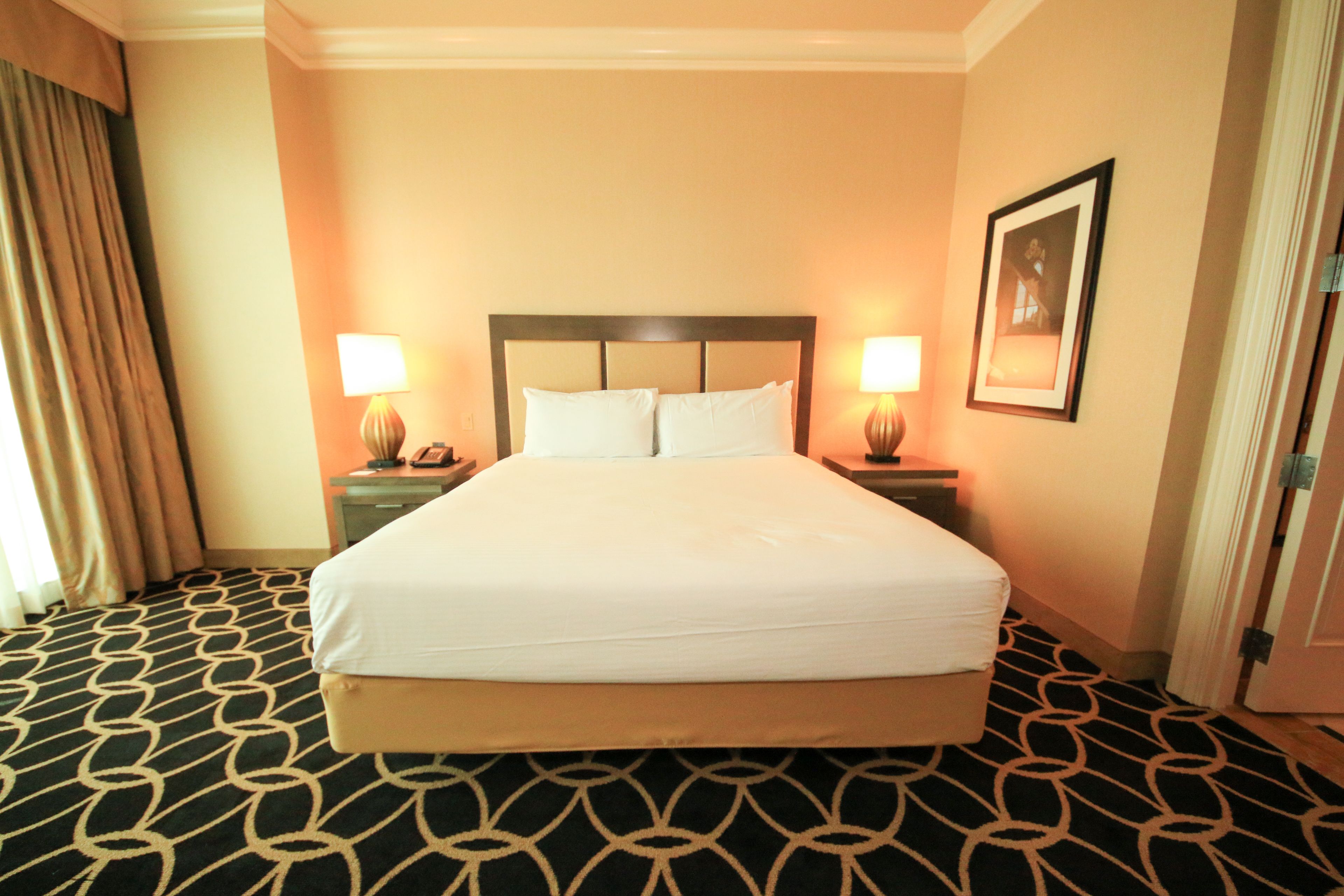 Habitación de hotel con una cama grande, dos mesitas de noche, una mesita auxiliar, un cuadro enmarcado en la pared y una alfombra estampada.