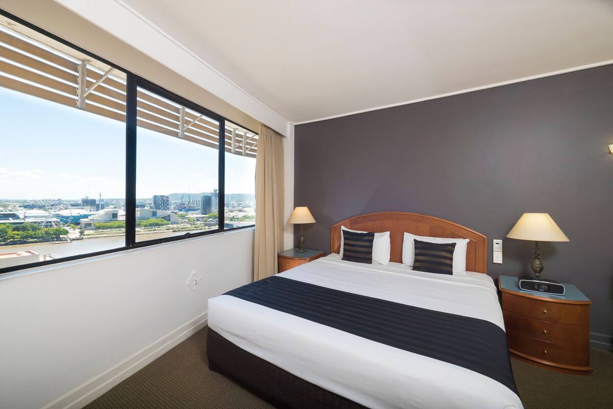 A hotel room with a large bed, two bedside tables, a window with blinds, and a view of a cityscape.