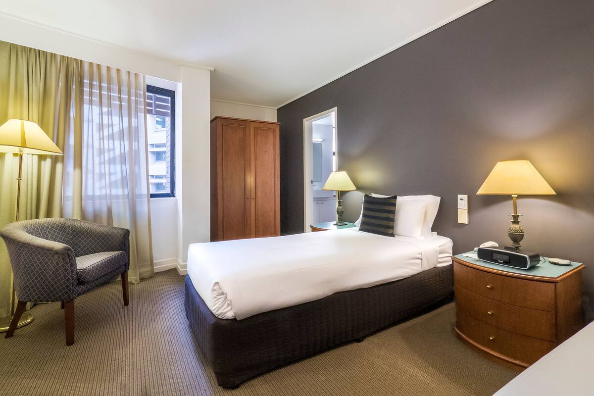 Ein Hotelzimmer mit einem Bett, zwei Nachttischlampen, einem Sessel, einem Schminktisch und einem Fenster mit Vorhängen.