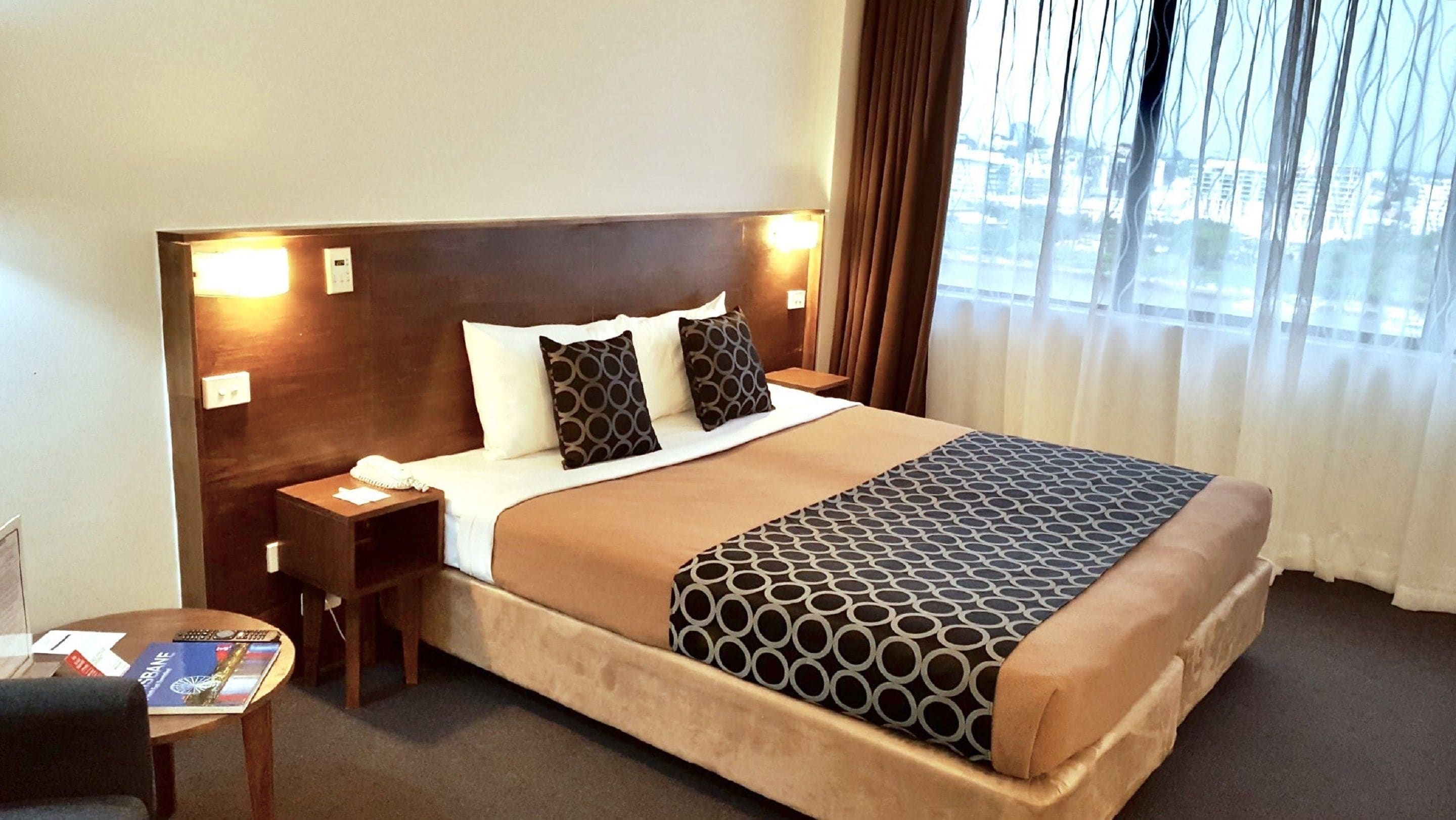 A hotel room with a large bed, a nightstand with a phone, a small table with a magazine, and a view of the city through the window.
