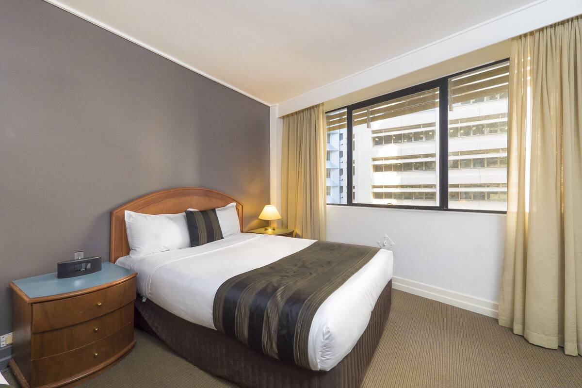 Ein Hotelzimmer mit einem großen Bett, einem Holfnachtkästchen, einer Lampe und einem Fenster mit Vorhängen.