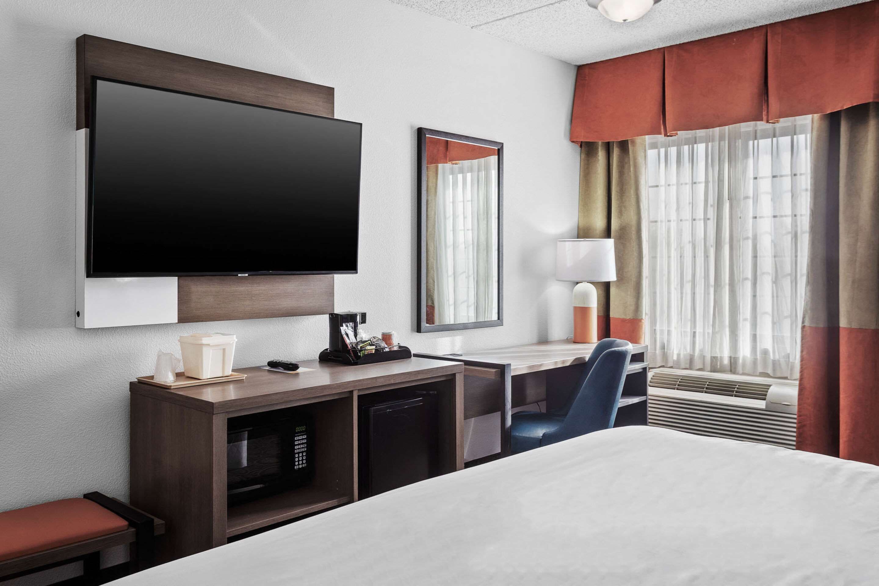 A hotel room with a large flat-screen TV, a desk with a lamp, a chair, a bed, a small kitchenette, and a window with curtains.