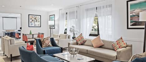 A modern living room with a sofa, armchairs, and a coffee table. There are two windows with sheer curtains, a painting on the wall, and a small table with a vase.
