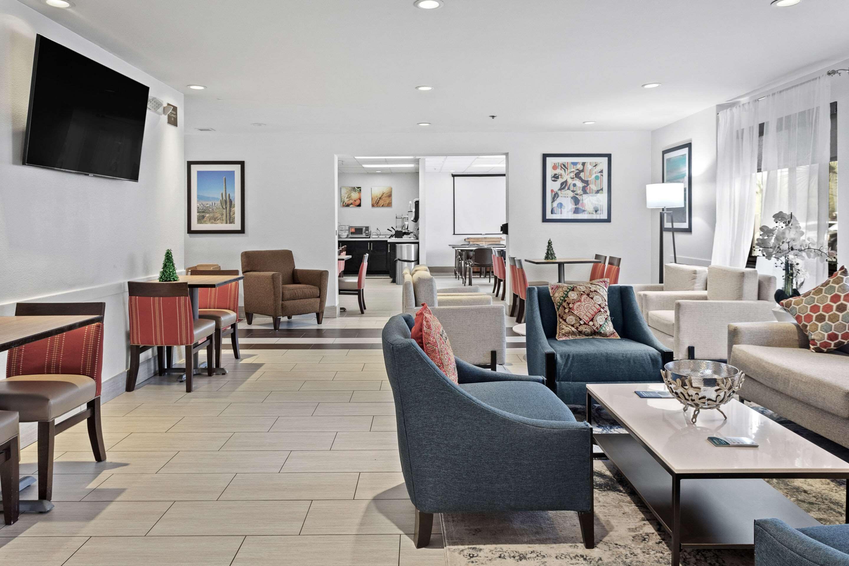 A modern hotel lobby with seating areas, a flat-screen TV, artwork, and a view into the kitchen area.