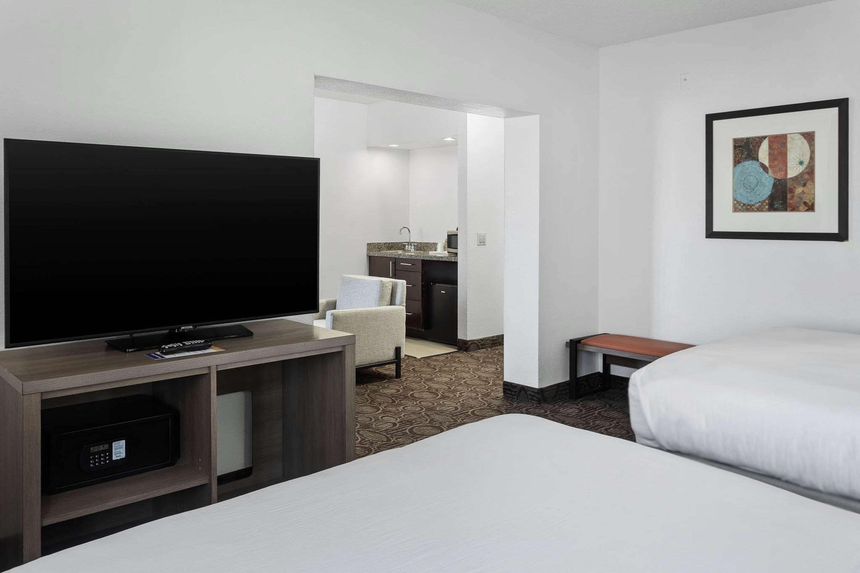A hotel room with a flat-screen TV, a bed, a small desk with a chair, and a kitchenette in the background.