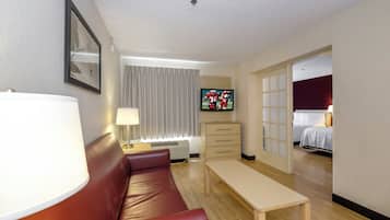 Quarto de hotel com sofá de couro vermelho, mesa de madeira, televisão, abajur e cama com roupa de cama branca.