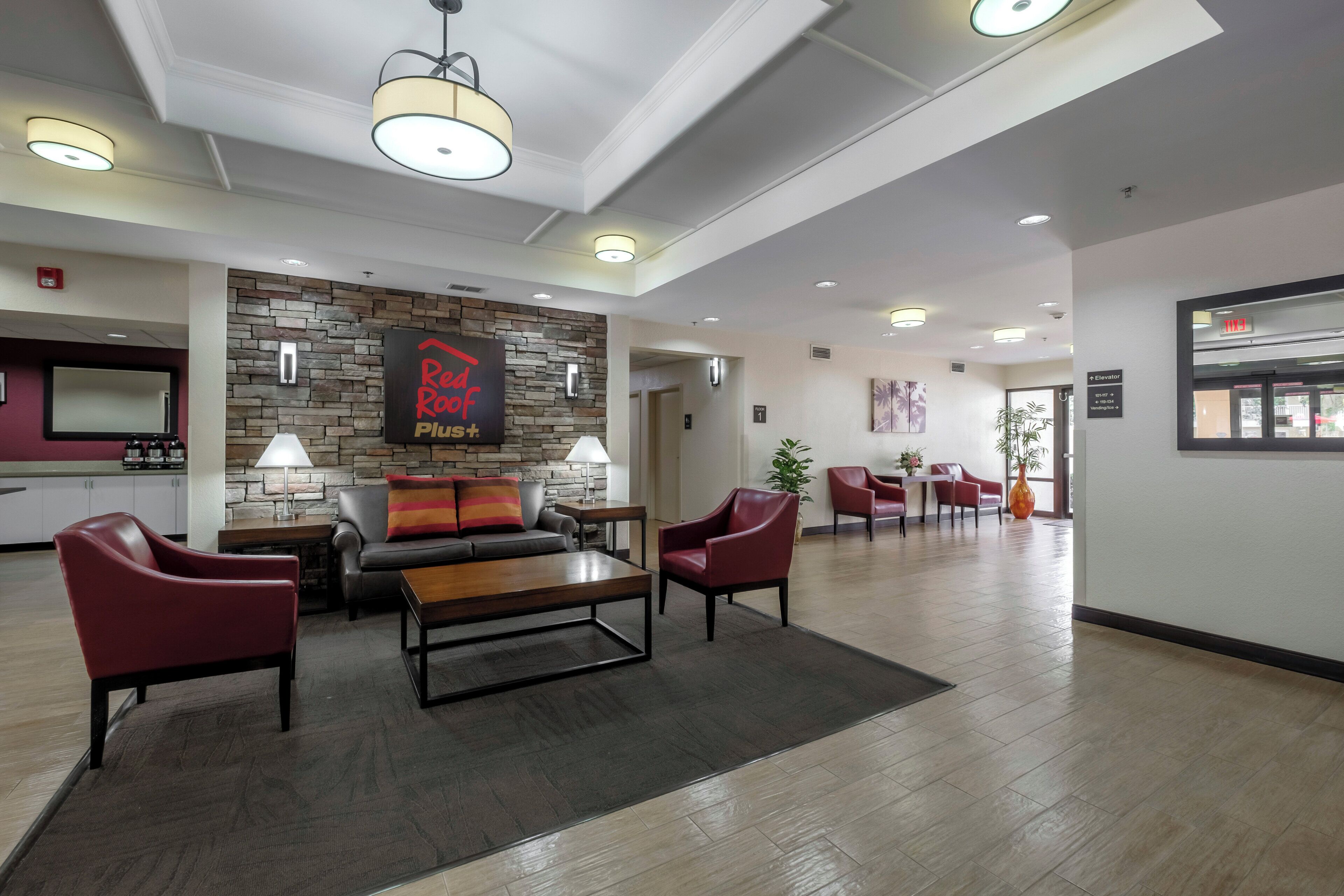 Un vestíbulo de hotel con pared de piedra, un letrero de 'Red Roof Plus', sillones de cuero, una mesa de centro de madera y un mostrador de recepción.