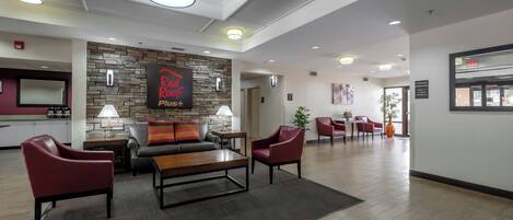 Un hall d’hôtel avec un mur en pierre, une enseigne « Red Roof Plus », des fauteuils en cuir, une table basse en bois et un comptoir de réception.