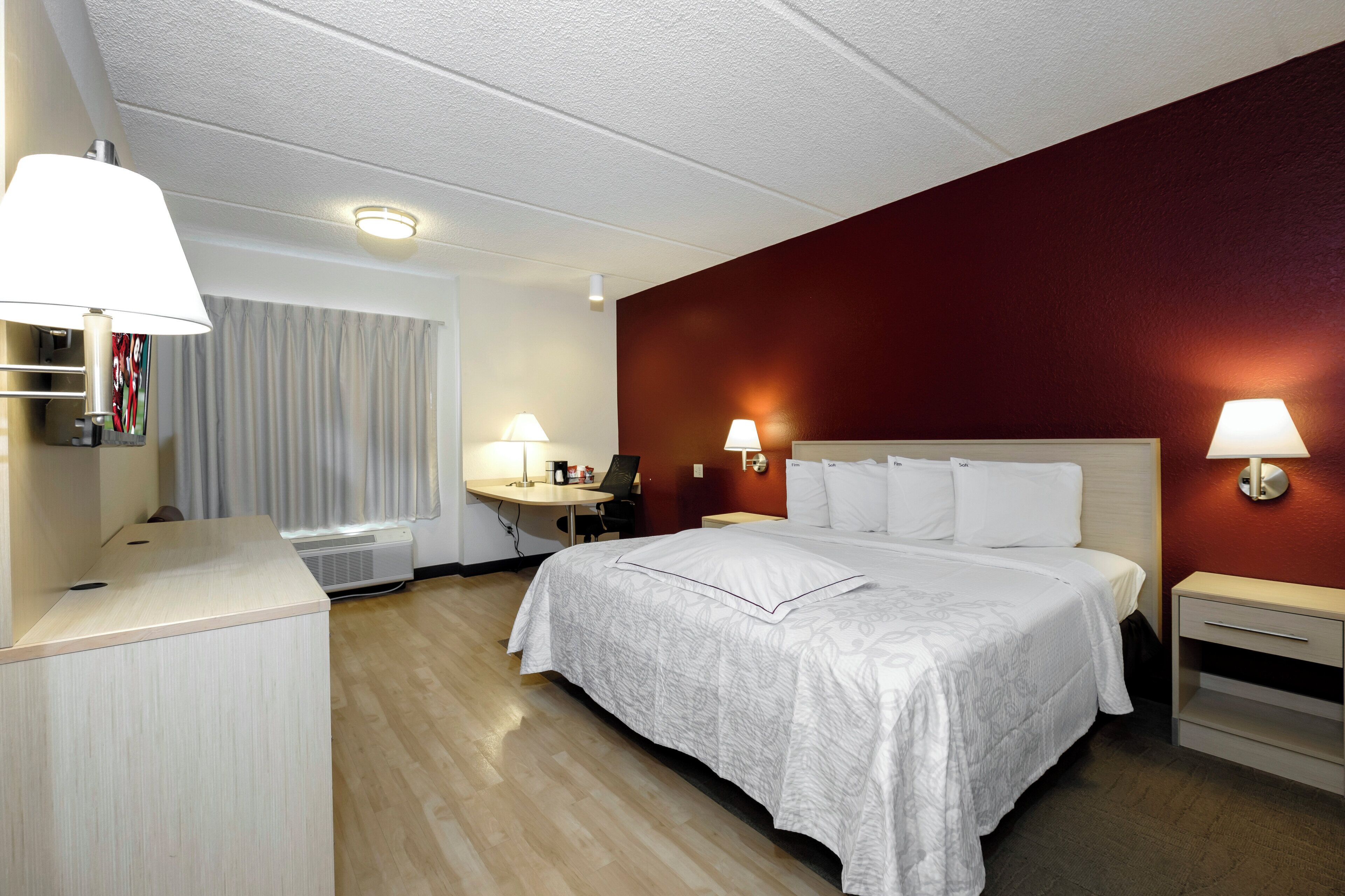 A hotel room with a bed, bedside tables, a desk, a chair, a lamp, a curtain, and a wall-mounted light fixture.