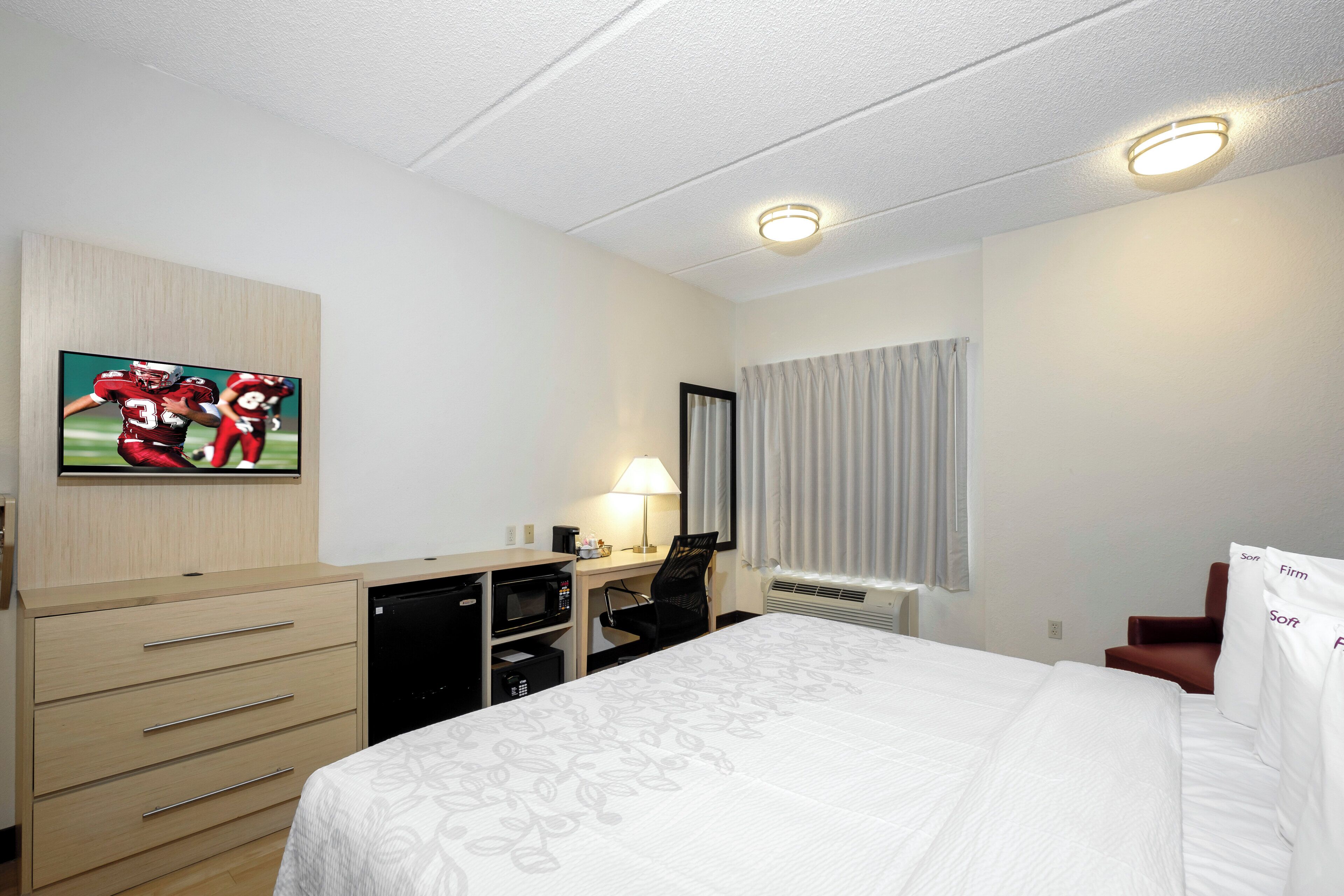 A hotel room with a bed, a desk, a chair, a TV, a lamp, a heater, and a window with curtains.