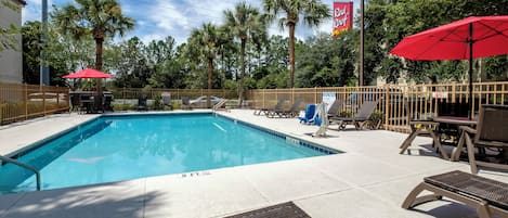 A swimming pool with lounge chairs and umbrellas, surrounded by palm trees and a fence.