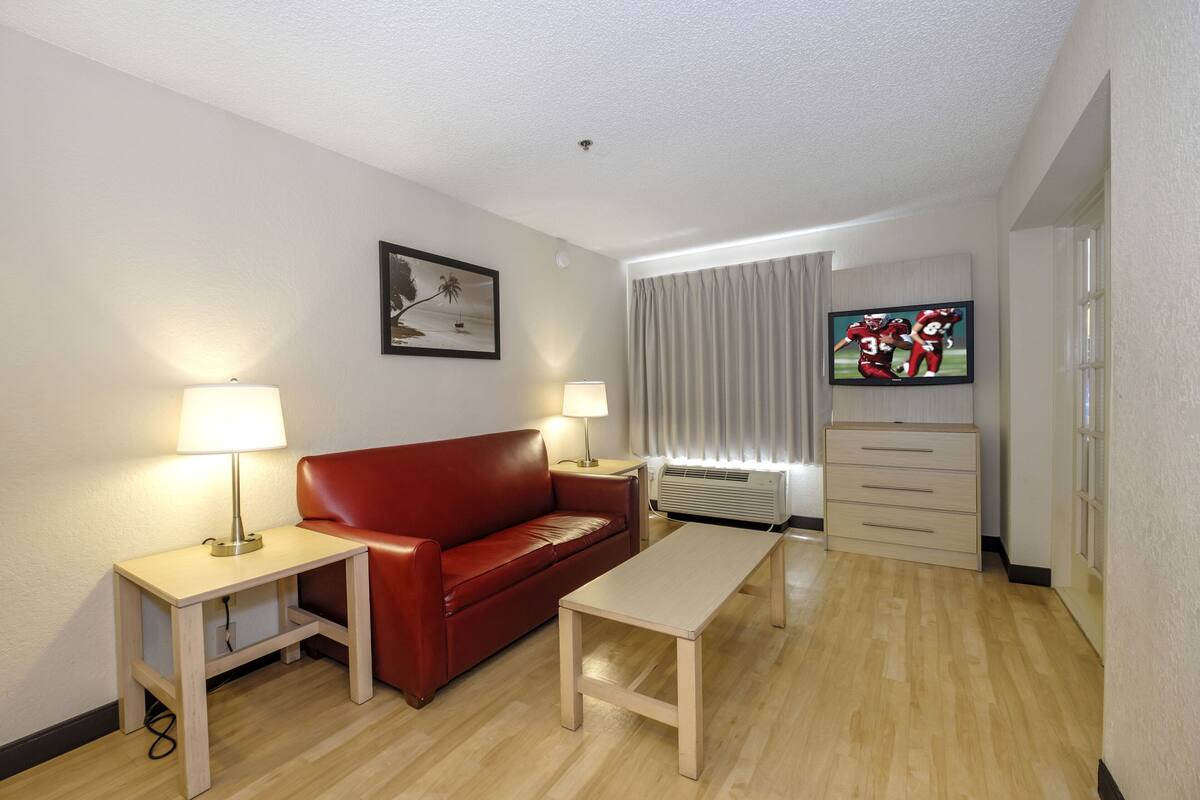 A hotel room with a red leather sofa, two bedside tables with lamps, a wooden coffee table, a TV on the wall, and a window with curtains.