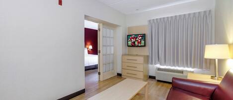 A hotel room with a wooden floor, a beige sofa, a small table, a TV mounted on the wall, a window with curtains, and a door leading to another room.