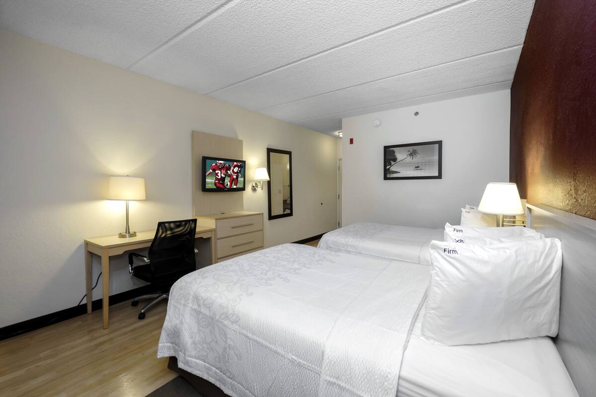 A hotel room with two beds, a desk, a chair, a TV, and a lamp.