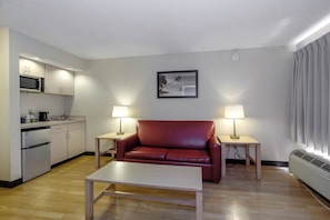 A compact living space with a kitchenette, a red leather sofa, two bedside lamps, and a framed picture on the wall. - Red Roof Inn PLUS+ Gainesville (Gainesville)