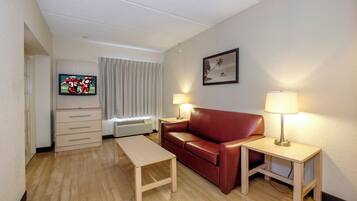 A hotel room with a television, a red leather sofa, a wooden table, two bedside lamps, a window with curtains, and a wall-mounted picture.