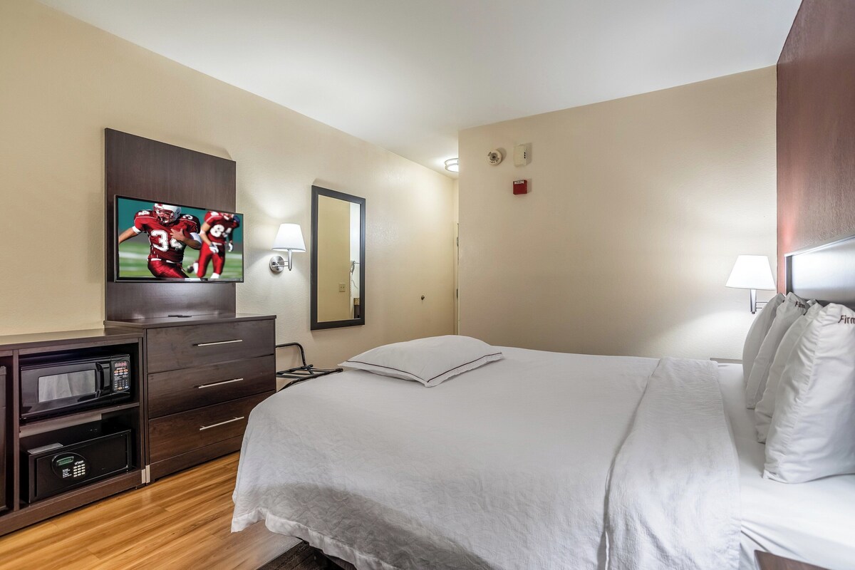 A hotel room with a bed, a TV mounted on a cabinet, a microwave, a mirror, and a lamp.