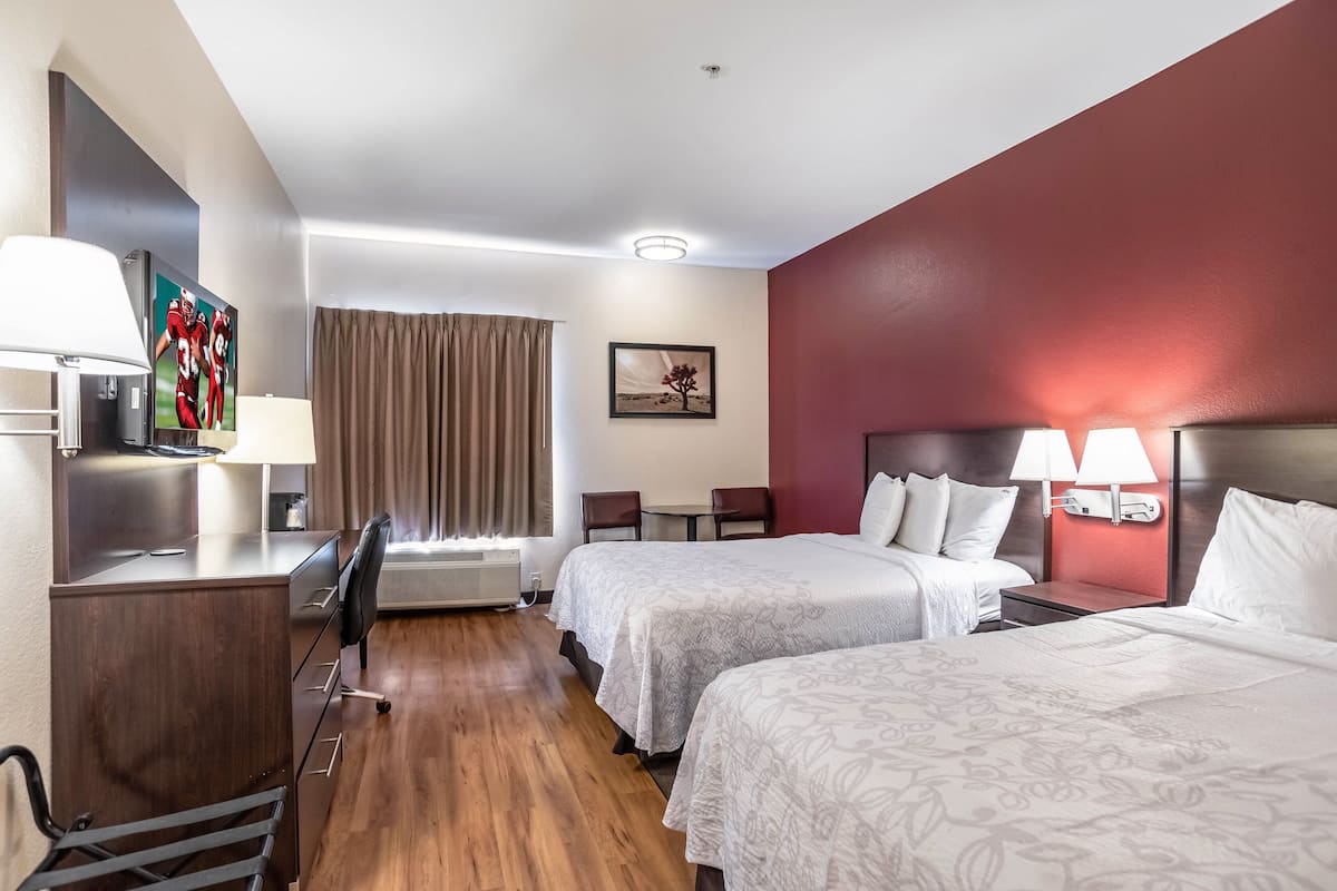 Ett hotellrum med två sängar, ett skrivbord med en lampa, en TV, en stol, ett litet bord och ett fönster med gardiner.