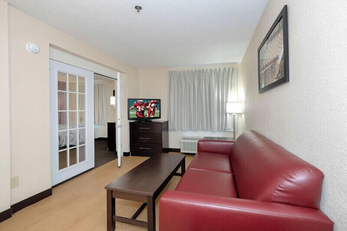 A living room with a red leather sofa, a wooden coffee table, a TV on a stand, a sliding glass door leading to a balcony, and a framed picture on the wall.