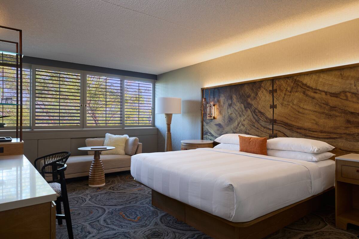 A hotel room with a large bed, a desk, a chair, a small table, and a lamp.