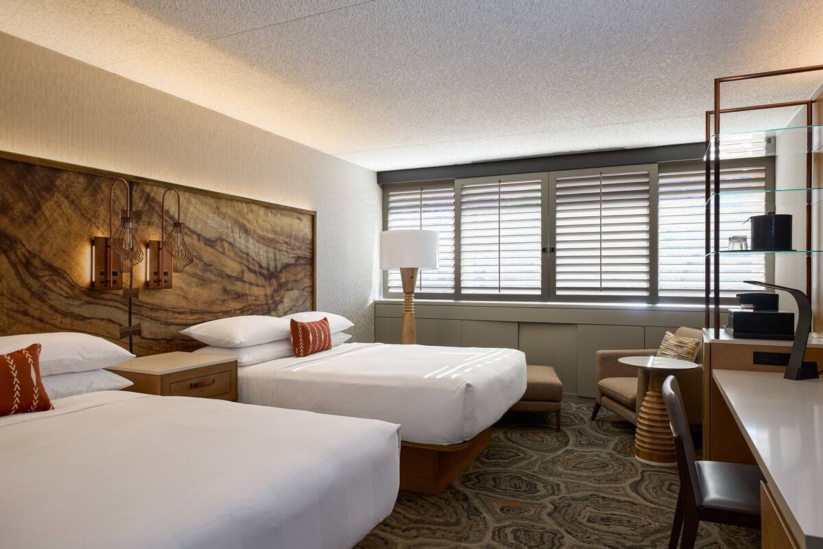 A hotel room with two beds, a desk, a chair, a lamp, and a window with blinds.