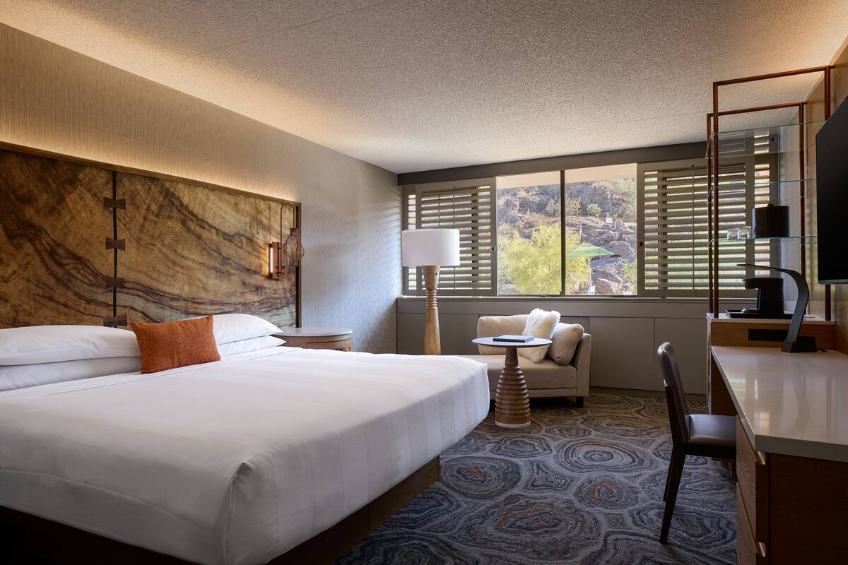 A hotel room with a large bed, a desk, a chair, a lamp, and a view of the outdoors.
