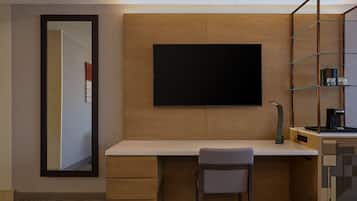 A modern hotel room with a wooden desk, a chair, a mounted flat-screen TV, and a glass shelving unit with a coffee maker.