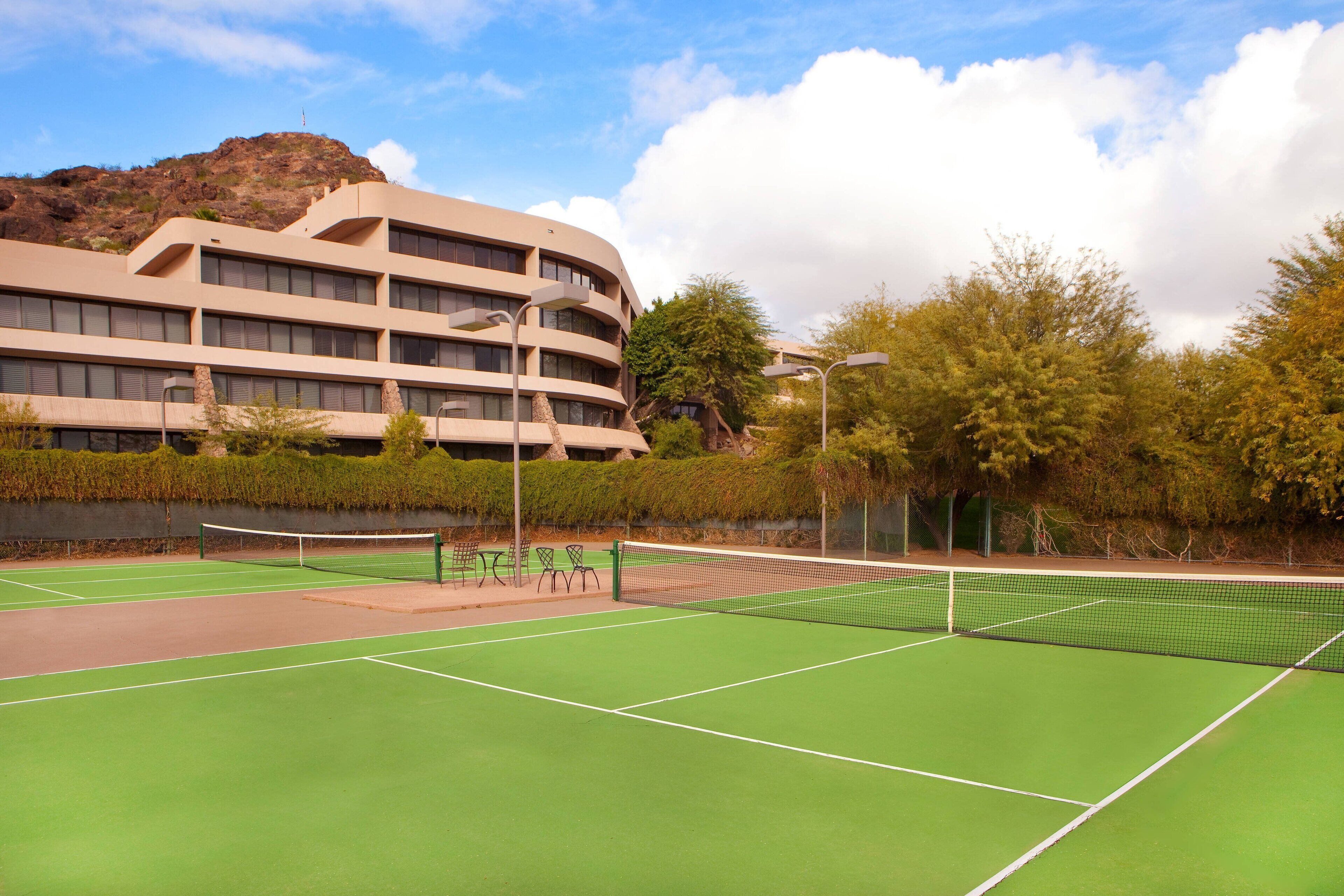 A tennis court with a net, surrounded by trees and a building with multiple floors.