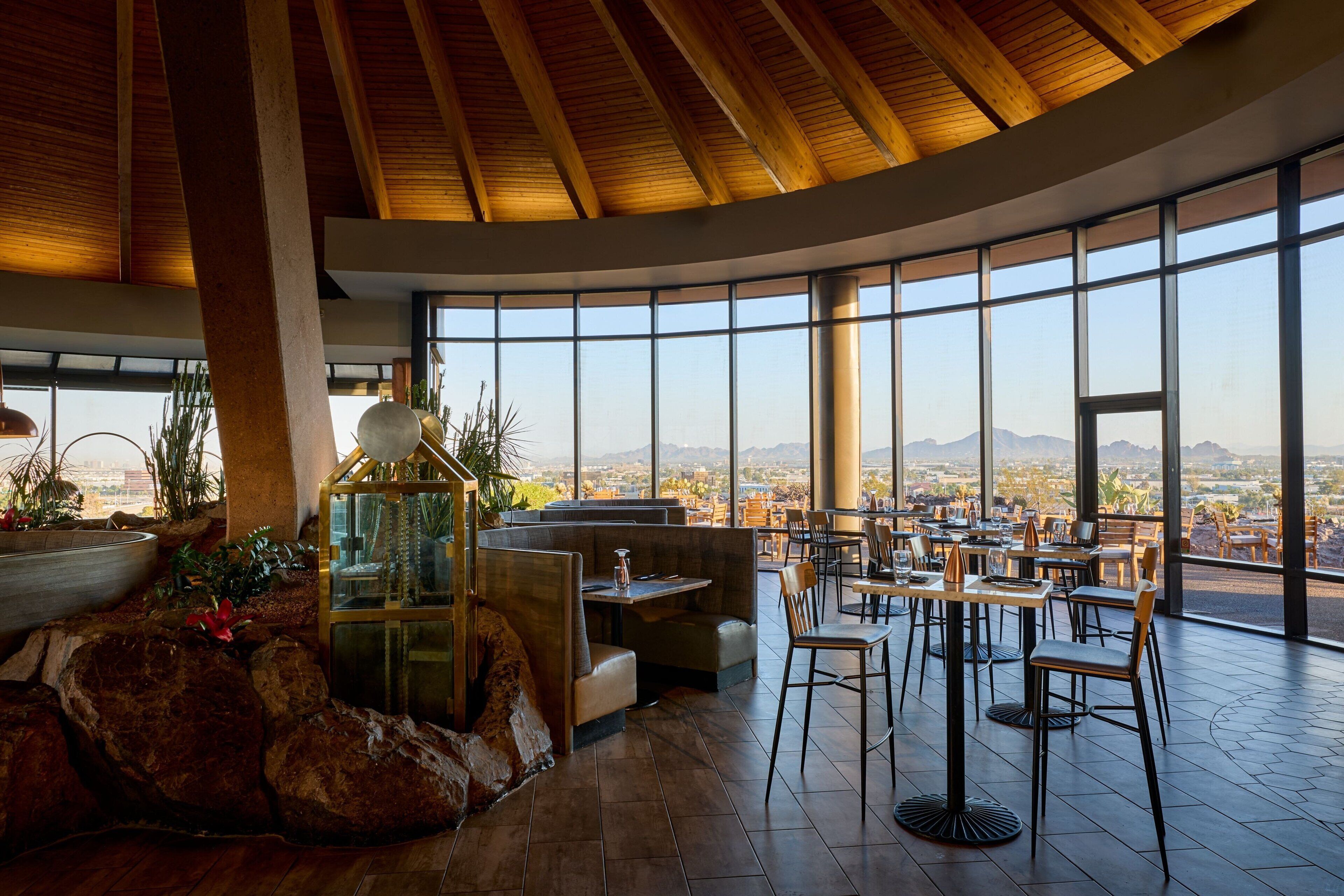 A restaurant with a view of the city and mountains, featuring wooden beams, large windows, and a mix of seating arrangements.