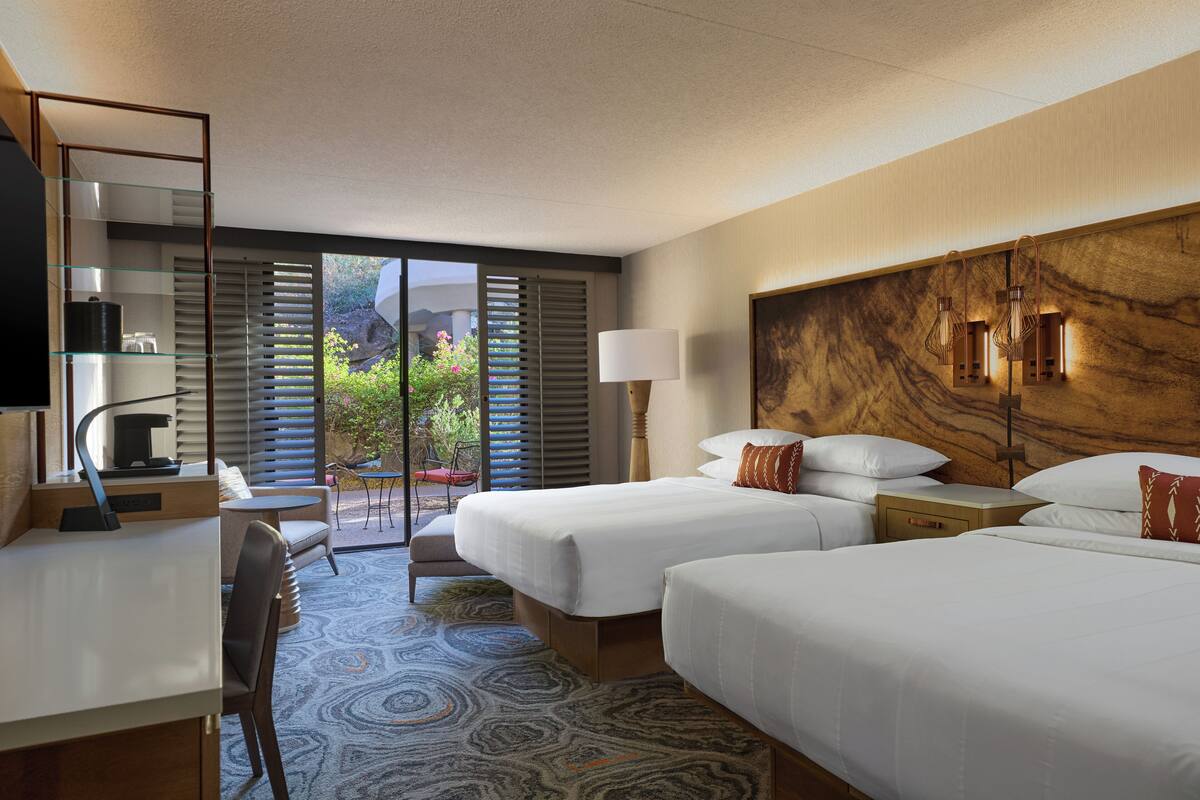 A hotel room with two beds, a desk, and a chair. There is a large window with blinds, a lamp, and a view of greenery outside.