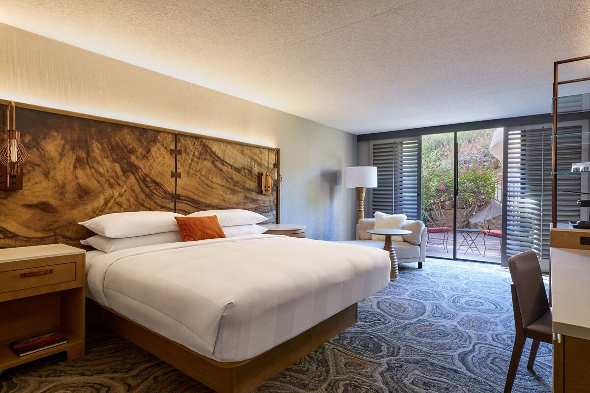 A hotel room with a large bed, a desk, a chair, a lamp, and a view of the outdoors.