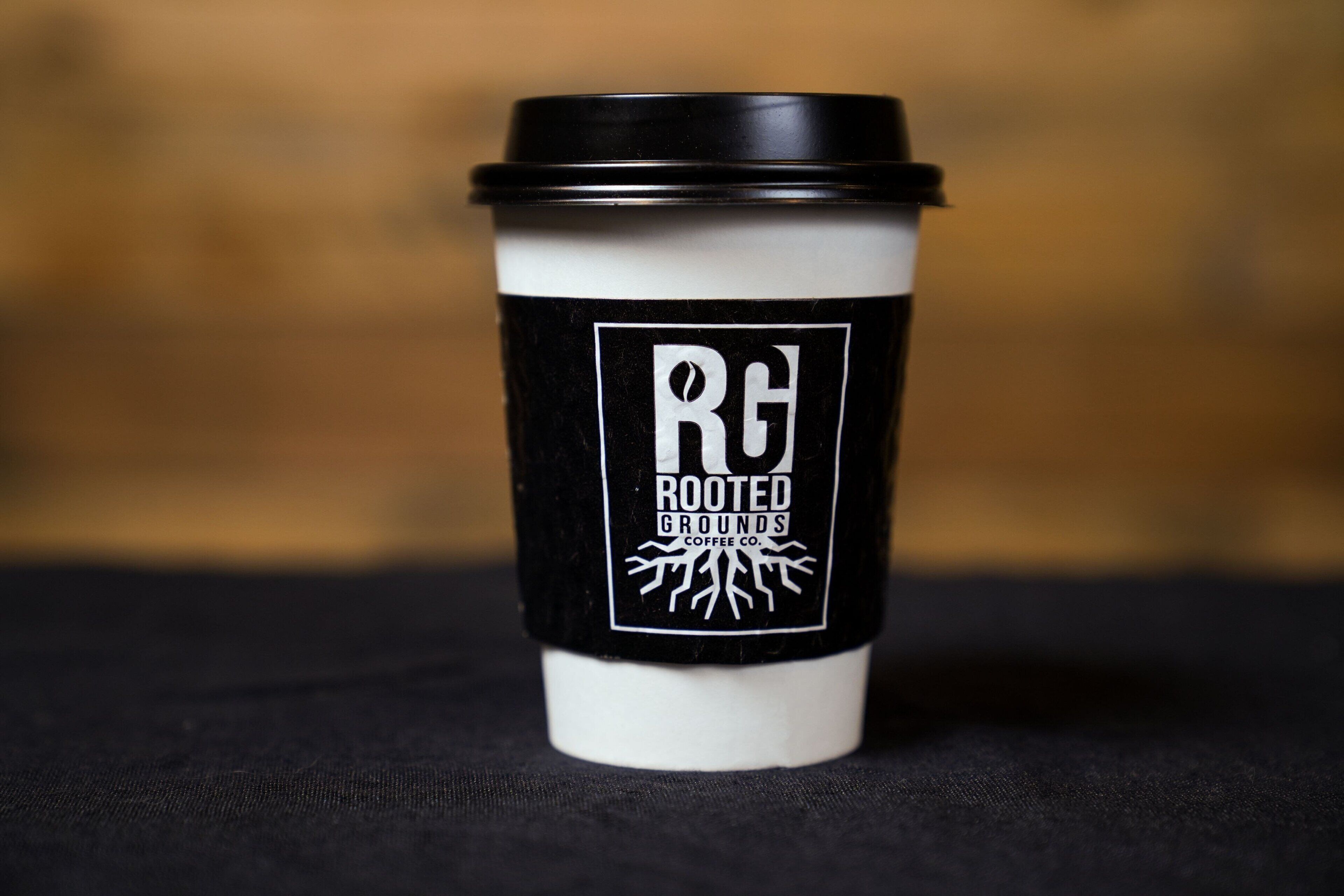 Uma xícara de café com tampa preta e capa branca, exibindo o logotipo e o nome 'Rooted Grounds Coffee Co'.