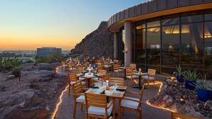 Una zona d'exterior per dinar amb taules i caires, un paisatge rocos i un edifici amb grans finestres.