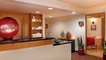 A reception area with a wooden reception desk, shelves with toiletries, a large red decorative piece, and potted plants.
