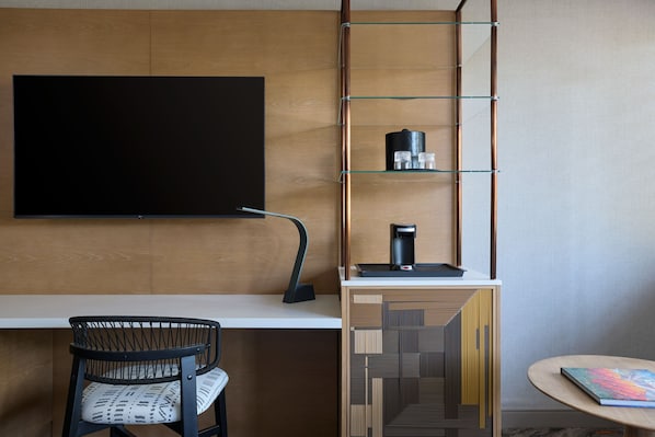 A modern room with a flat-screen TV mounted on the wall, a coffee machine on a shelf, a patterned chair, and a small round table with a book.