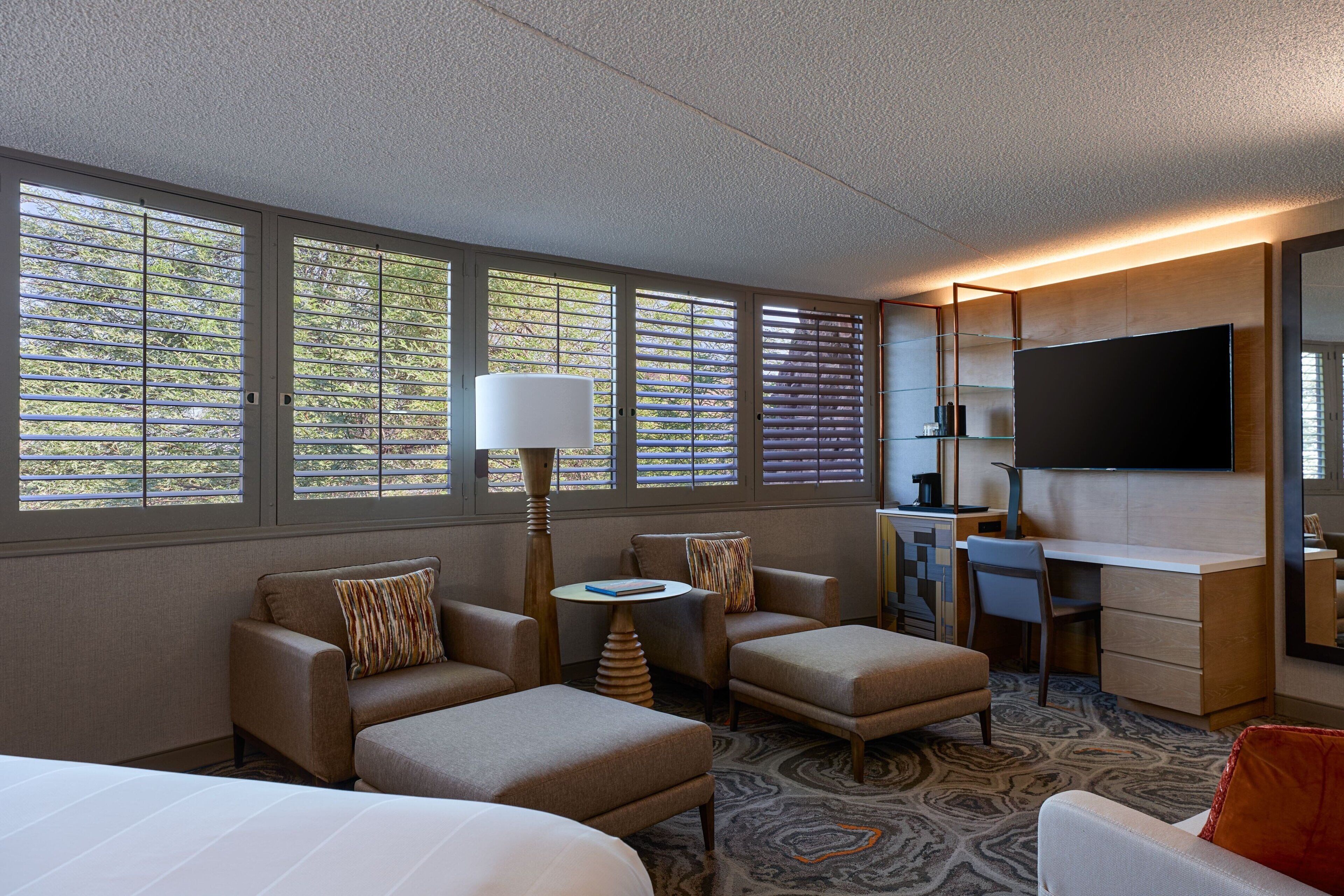 A hotel room with a bed, two armchairs, a desk, a television, and large windows with blinds.