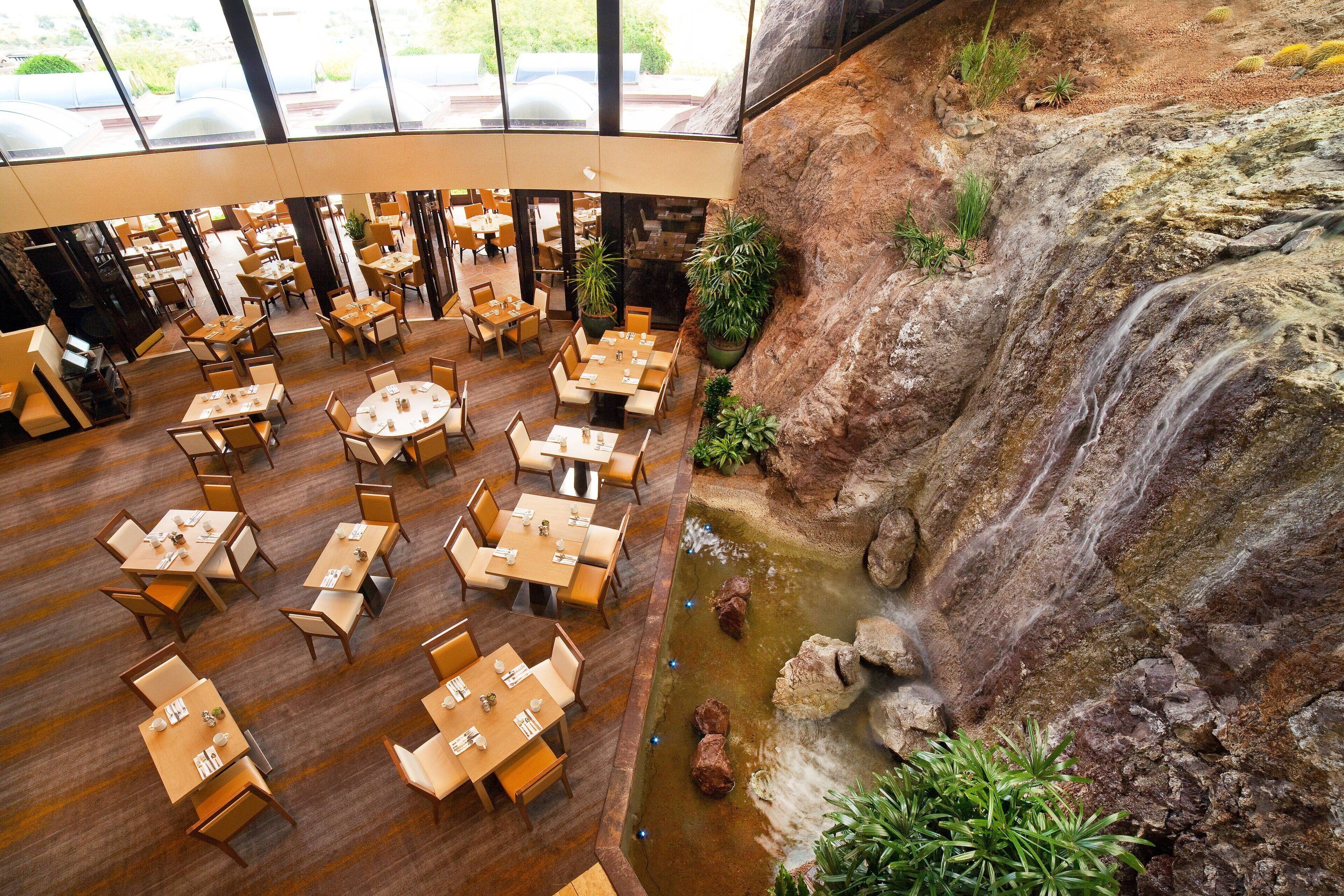 A spacious indoor dining area with multiple tables and chairs, a waterfall feature, and a view of the outdoors through large windows.