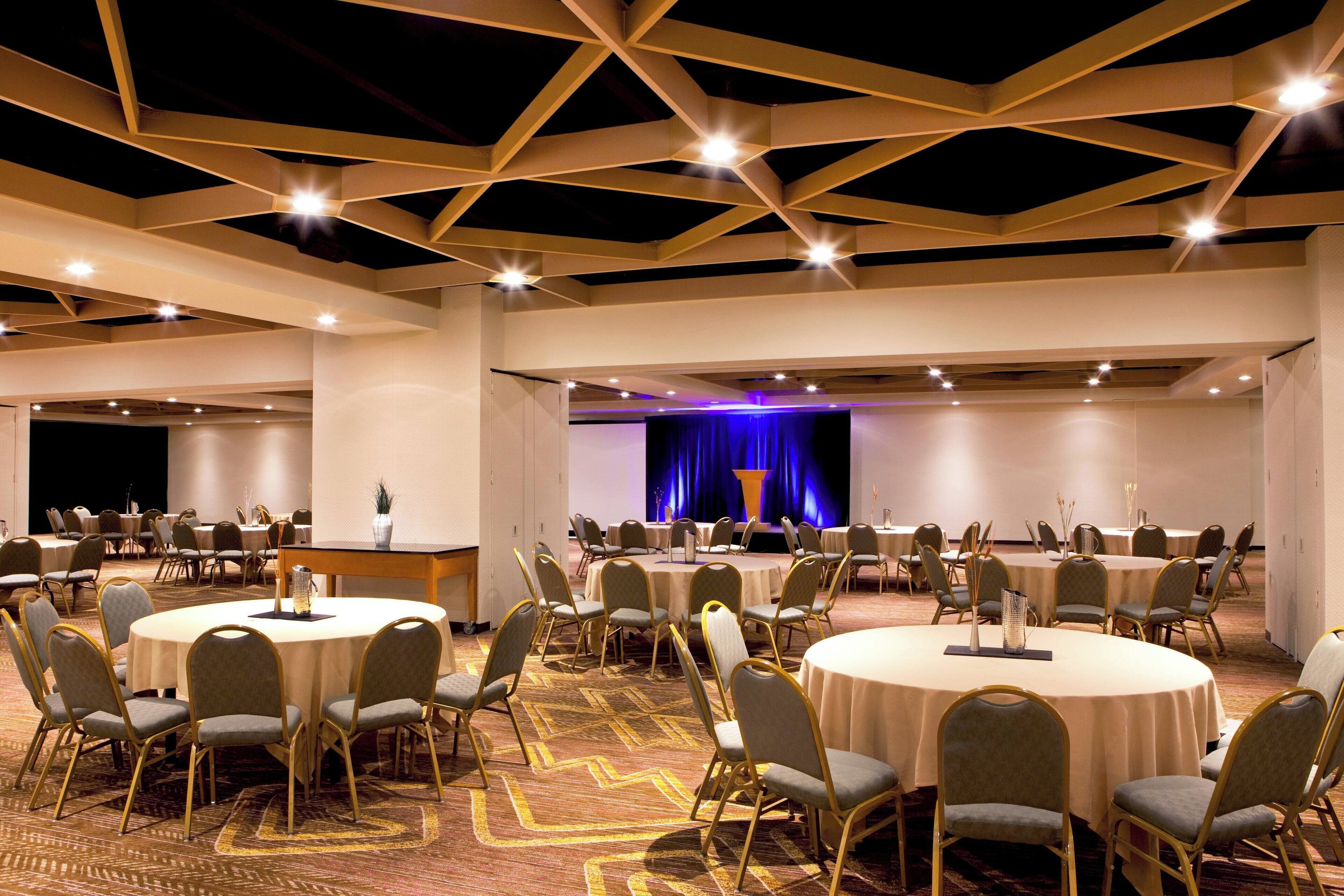 Une salle de conférence avec des tables et des chaises rondes, une scène avec un rideau bleu et un tapis à motifs.