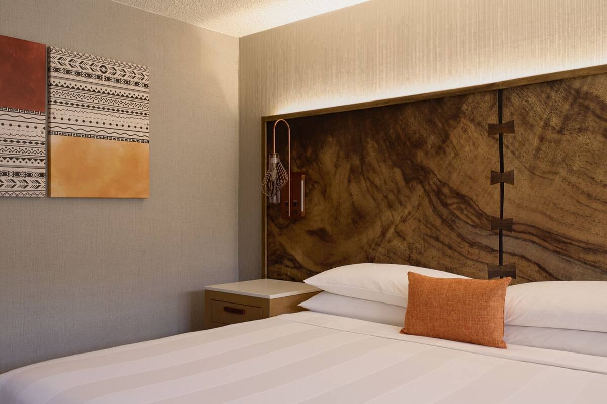 A hotel room with a wooden headboard, a bed with white linens, a nightstand, and a wall with patterned artwork.