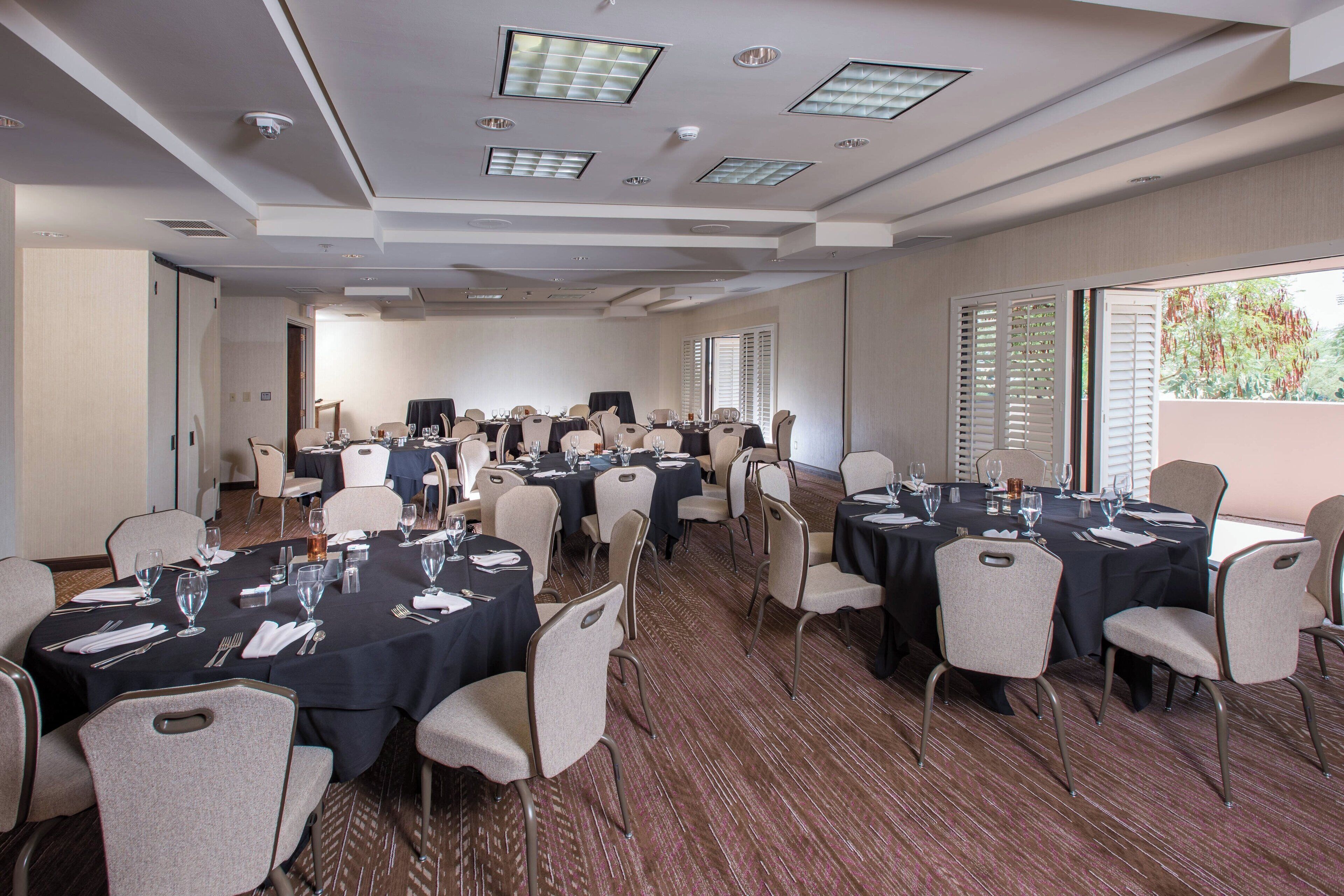A conference room with multiple tables and chairs, equipped with glassware and linens, and large windows with blinds.