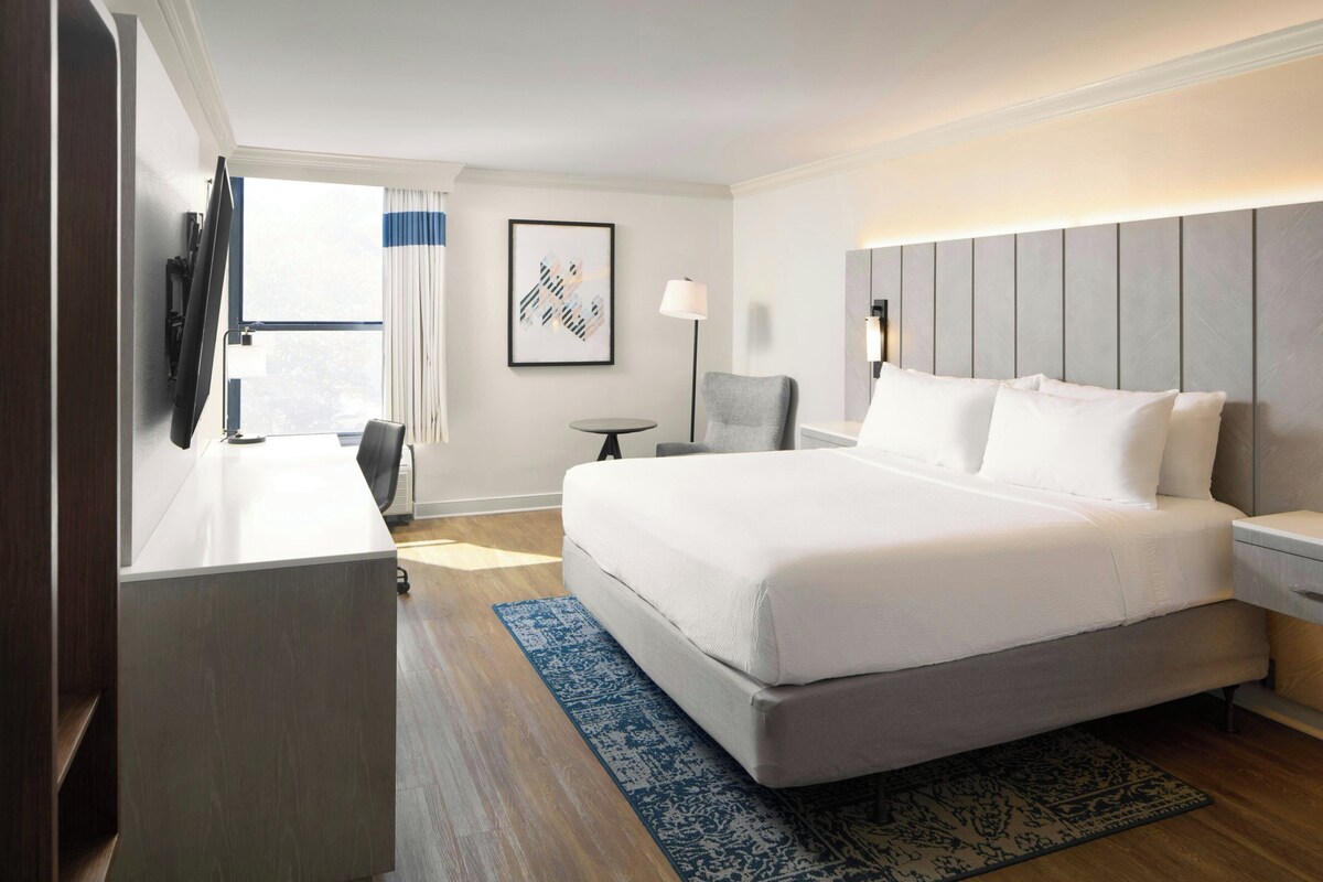 Een hotelkamer met een bed, een bureau, een stoel, een televisie, een lamp en een raam met gordijnen.