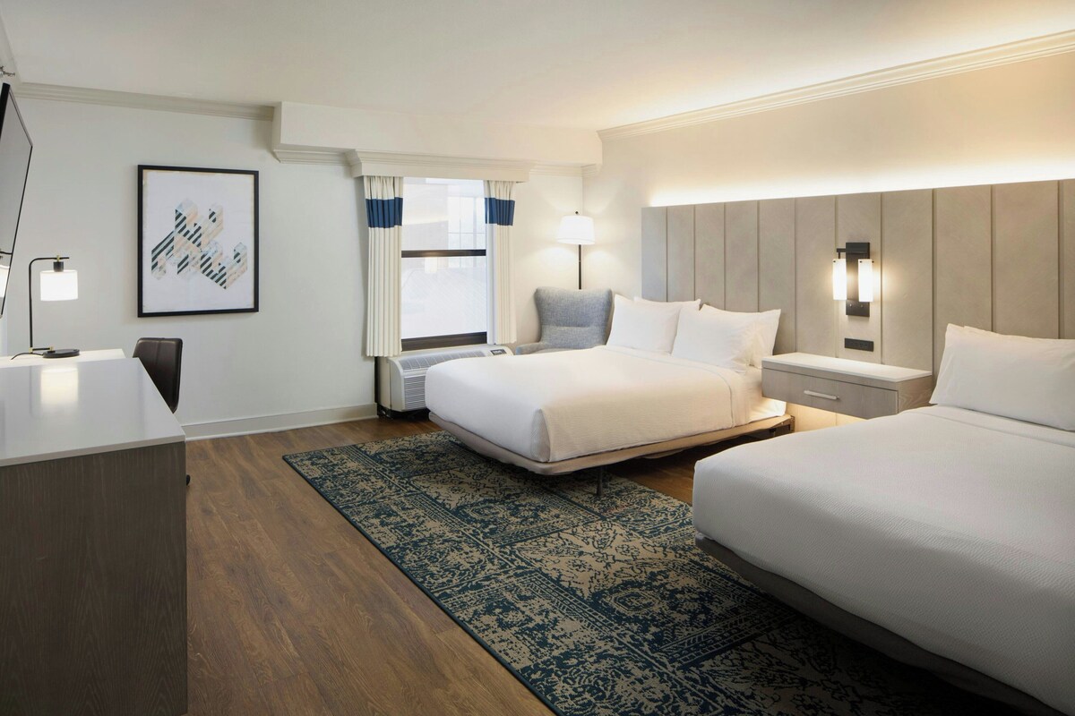 Een hotelkamer met twee bedden, een bureau, een stoel, een groot raam met gordijnen, een tv aan de muur en een lijst met een afbeelding.