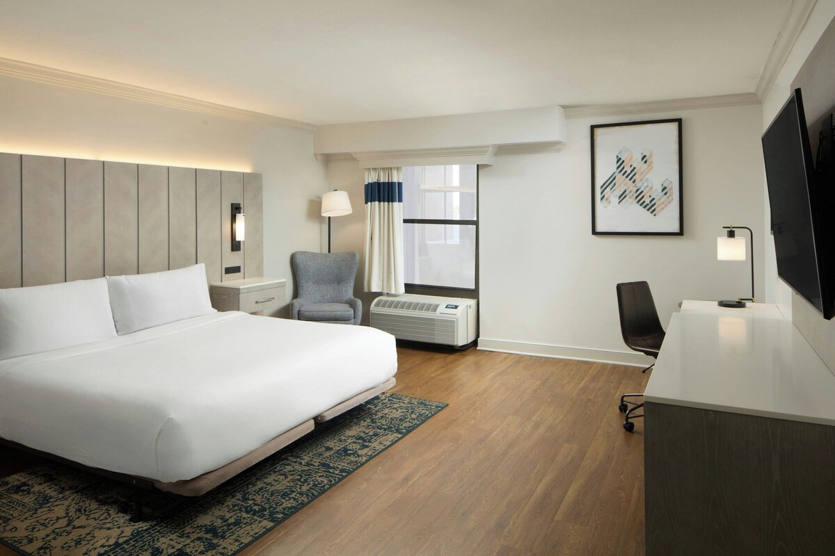 Een hotelkamer met een groot bed, een bureau, een stoel, een televisie, een raam met gordijnen en een kunstwerk aan de muur.